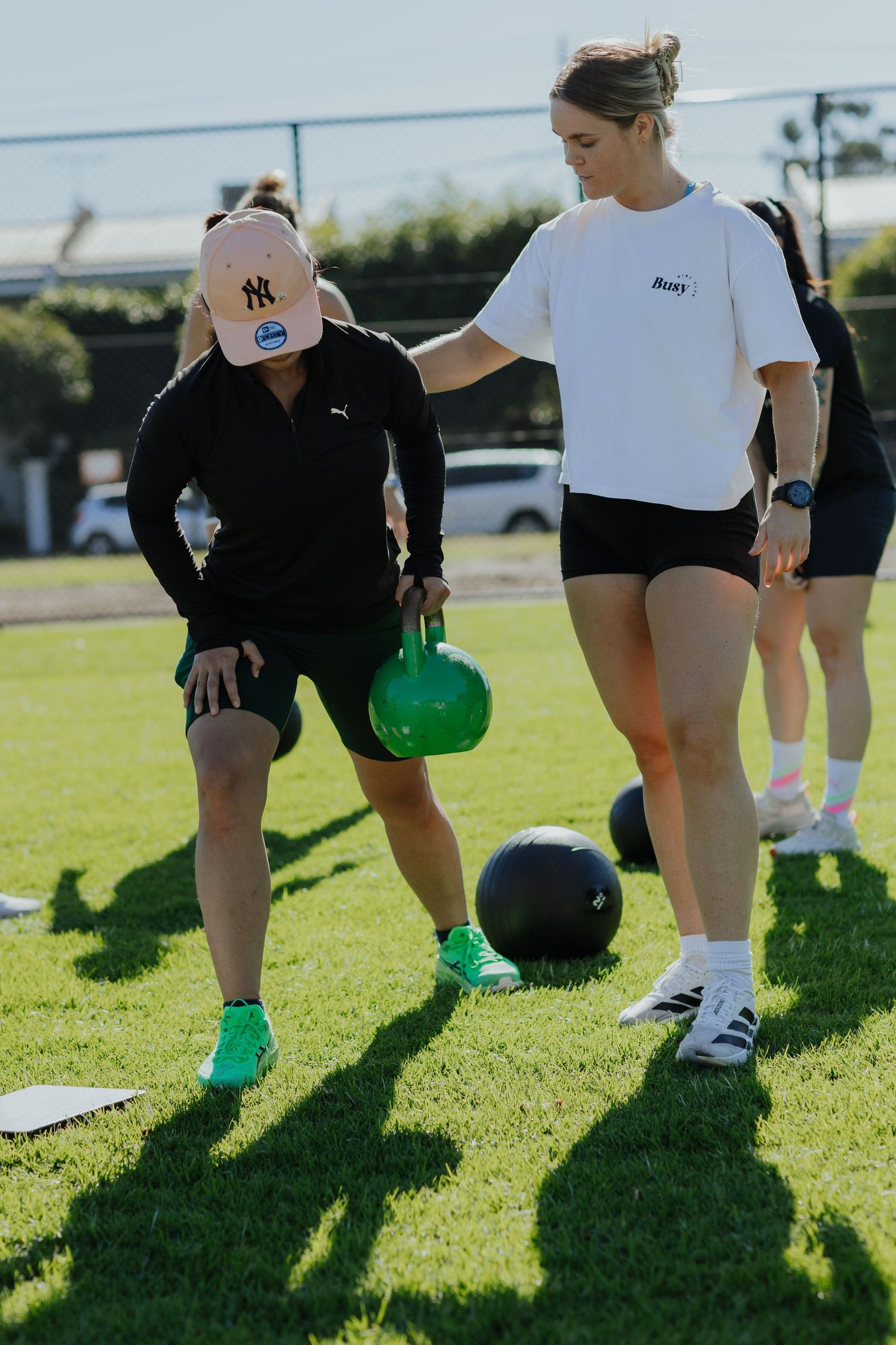Woman performing a kettlebell exercise on grass while a trainer in a Roxy shirt provides guidance.