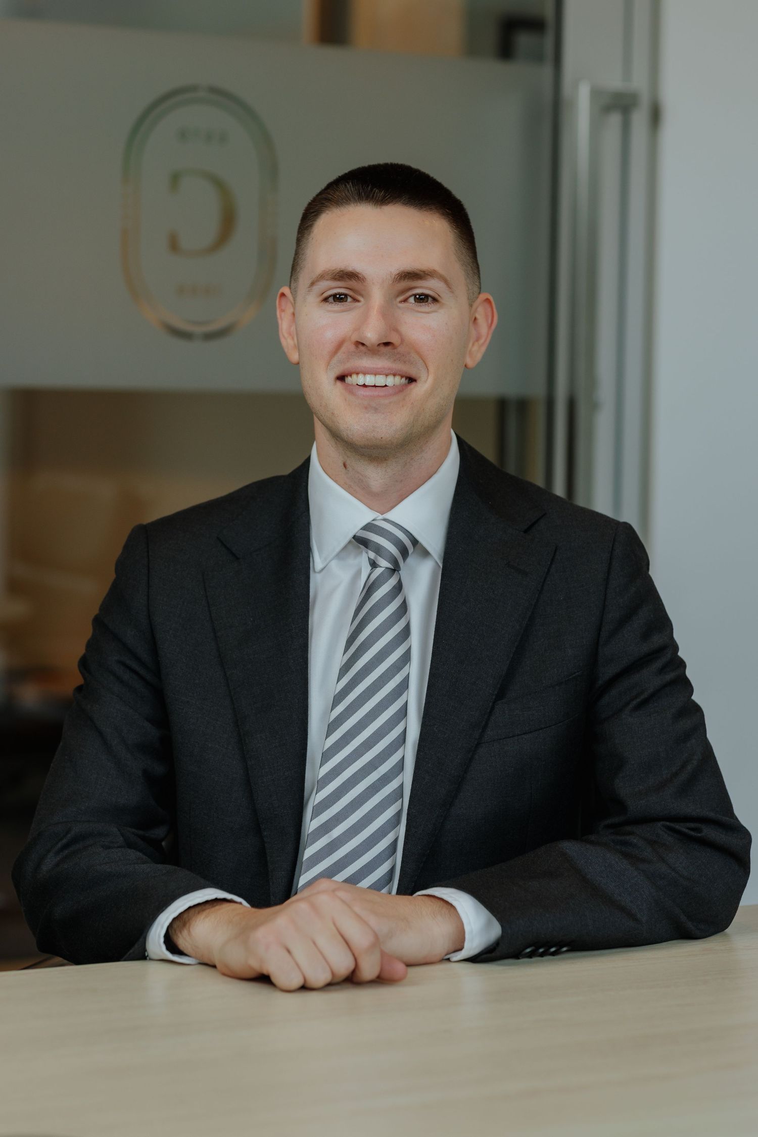 Smiling young businessman in dark suit and striped tie seated at an office desk.