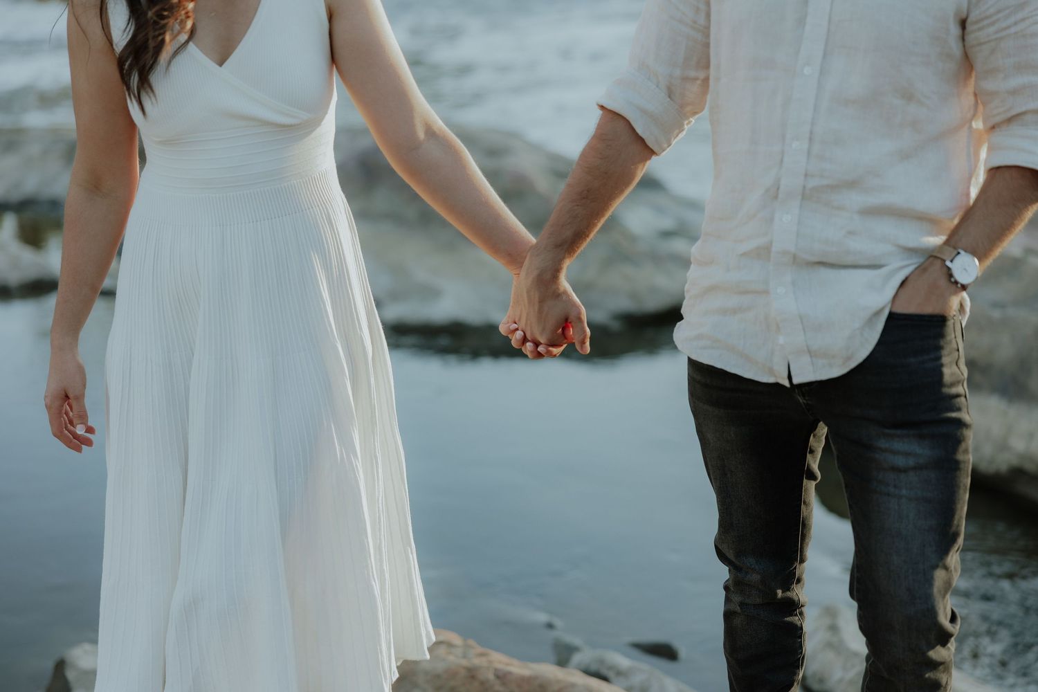Close-up of couple holding hands in white dress and dark jeans standing on rocks near water.