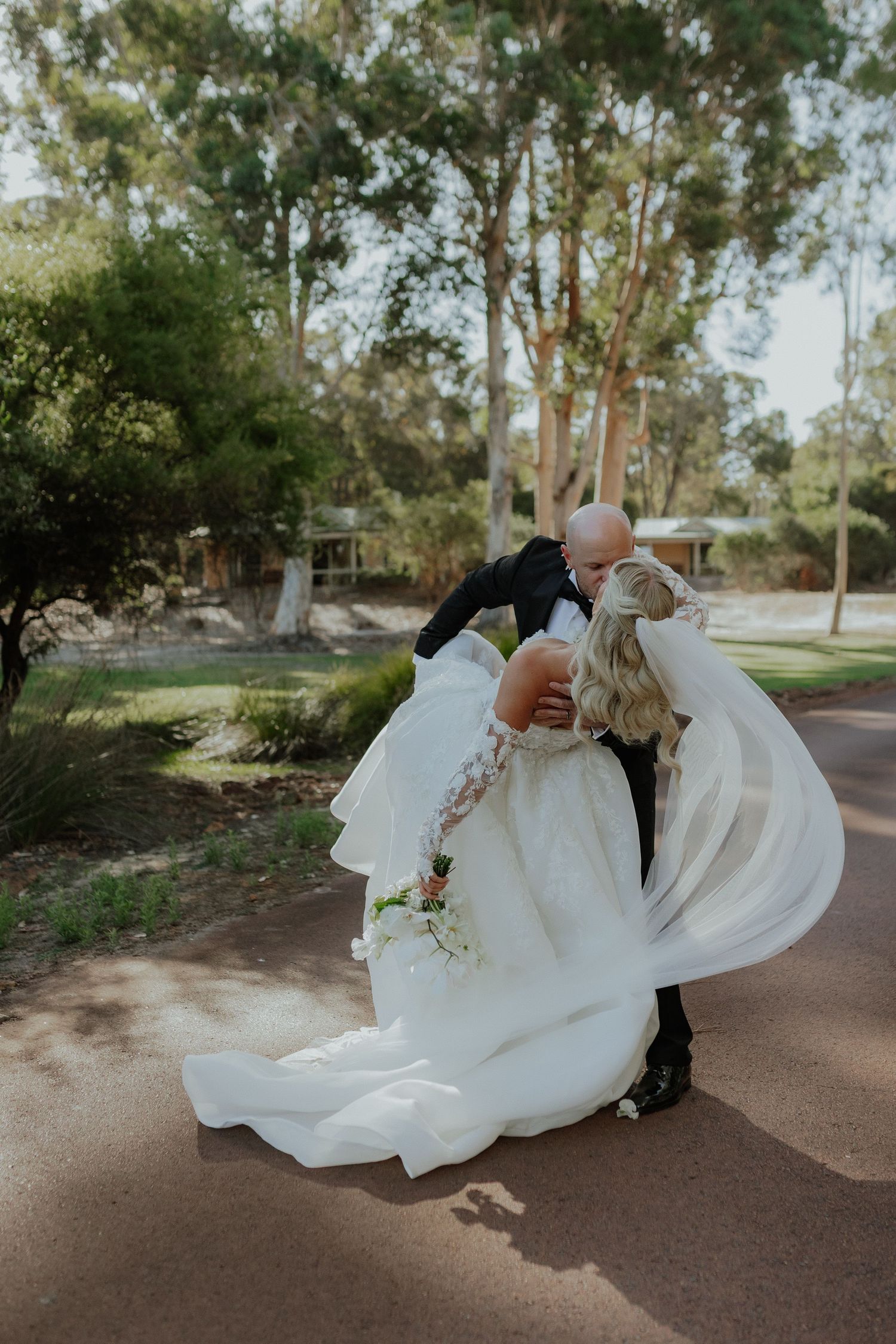 Groom dipping bride in a flowing white gown for a romantic kiss on a tree-lined path.