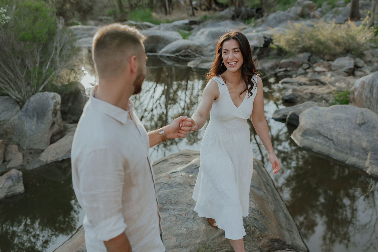 Smiling woman in white dress holds hands with man in white shirt near a rocky creek in nature.