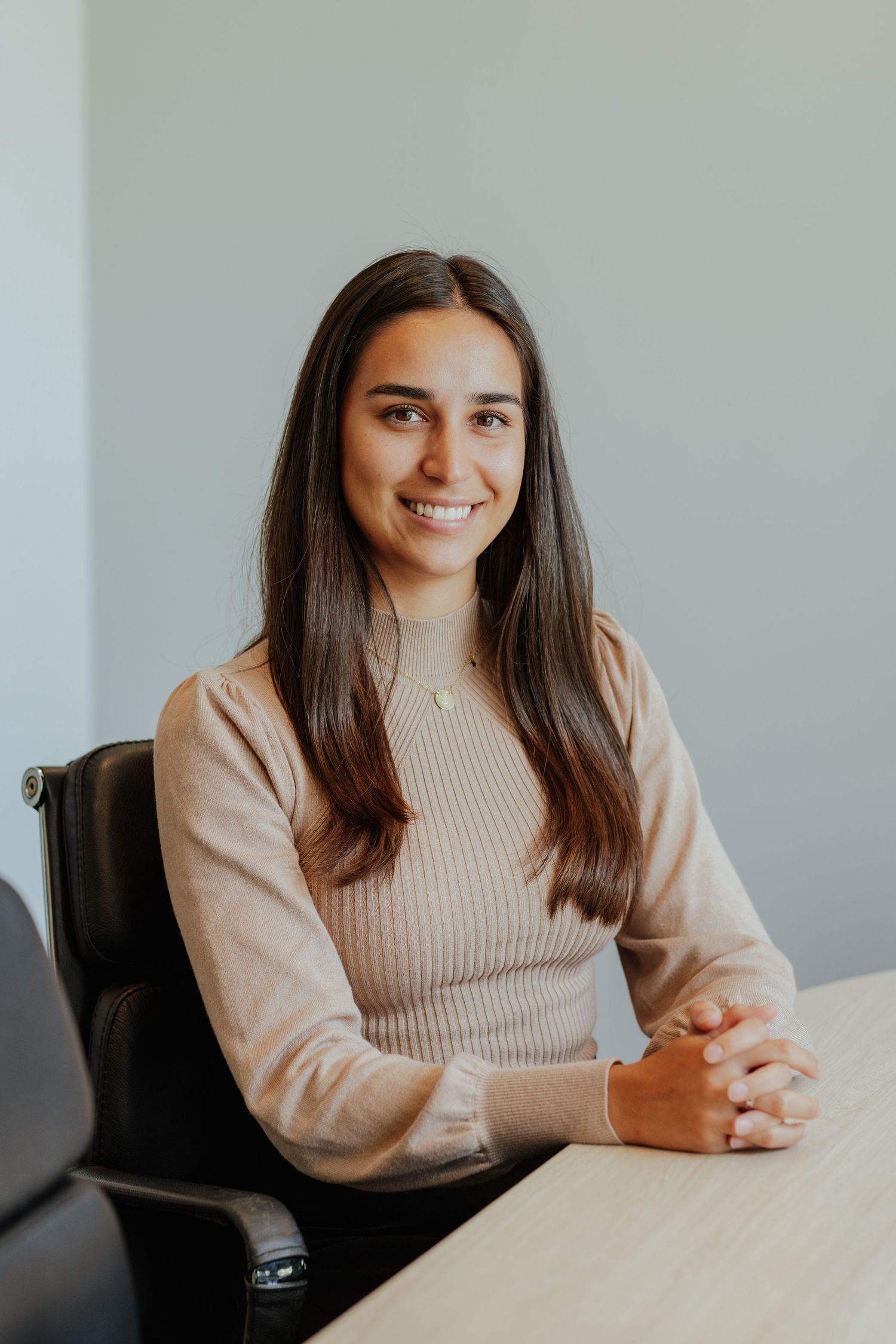 Young professional woman in beige sweater smiling with hands clasped at a desk.