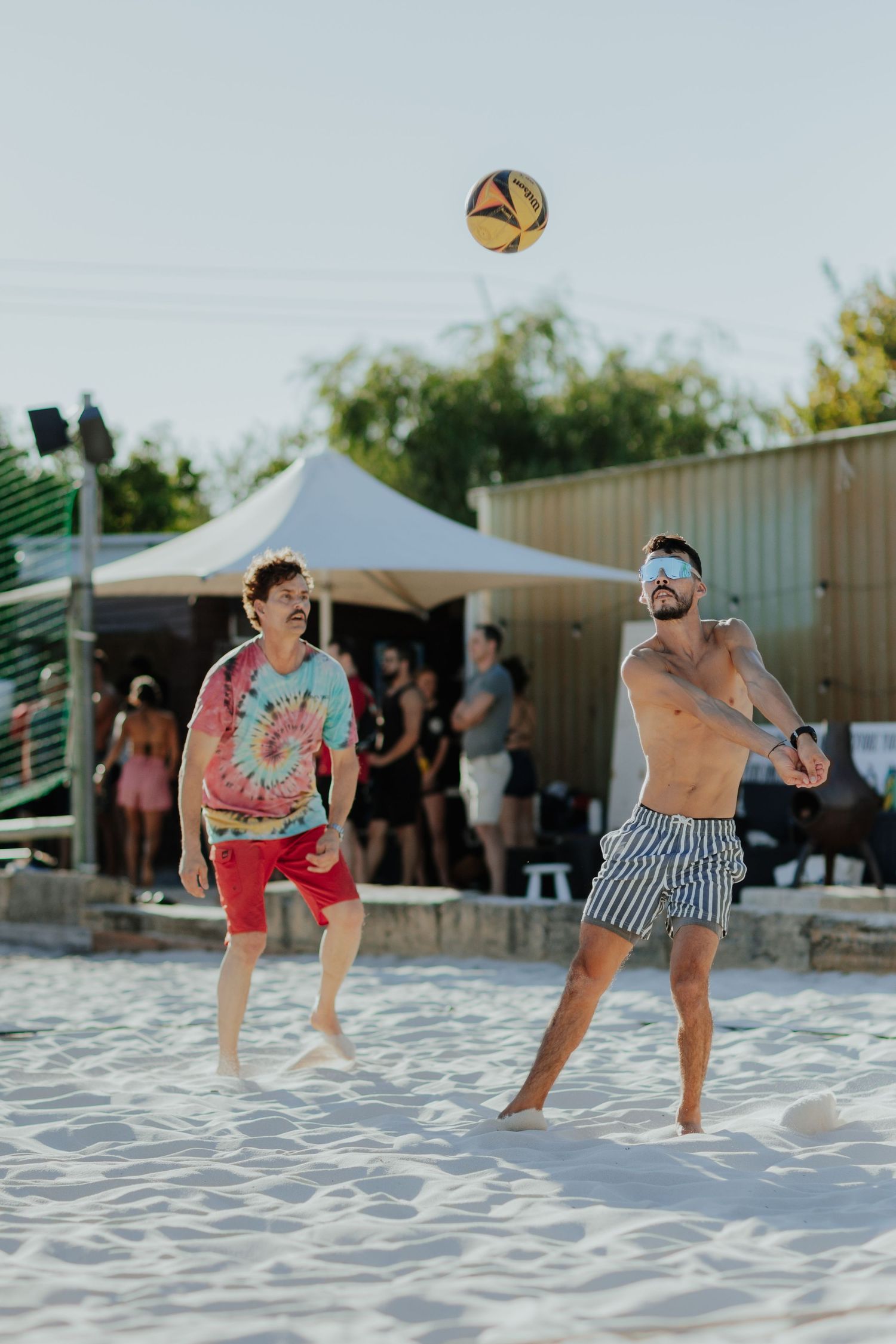 Two men playing beach volleyball on white sand, one shirtless in striped shorts, the other in a tie-dye shirt and red shorts.