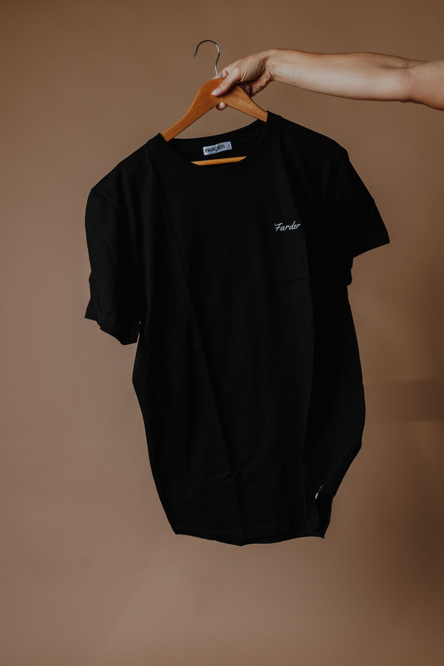 Black Farder branded t-shirt displayed on a wooden hanger against a warm beige background.