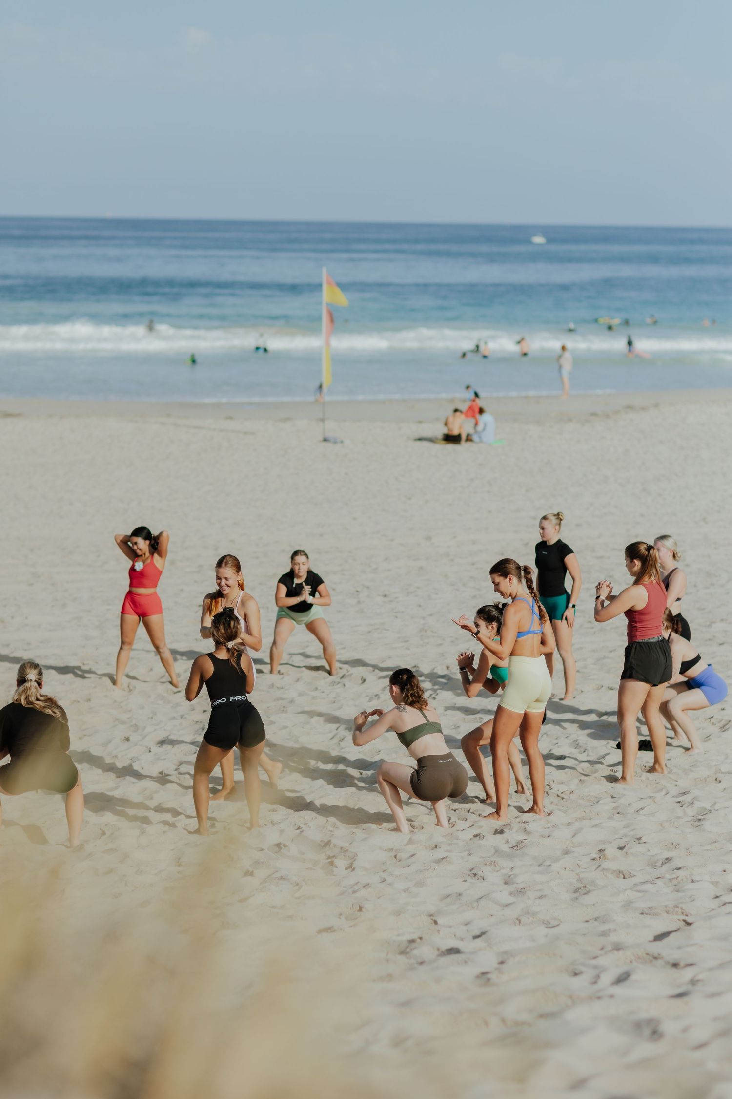 Group of women in athletic wear exercise together on a sunny sandy beach near the ocean.