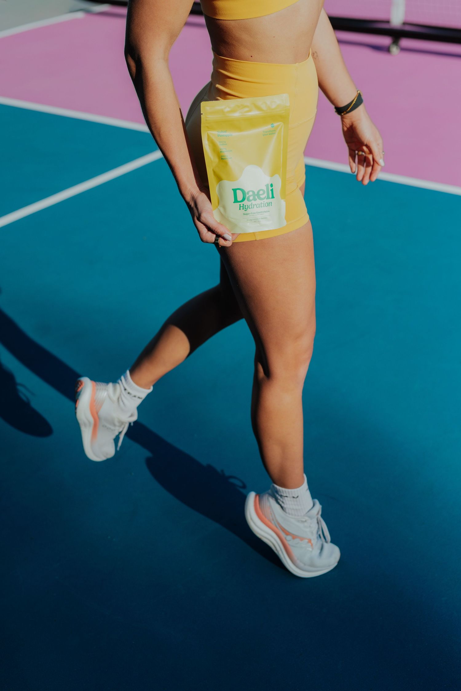 Woman in yellow activewear holding a Daeli Hydration pouch while walking on a tennis court.