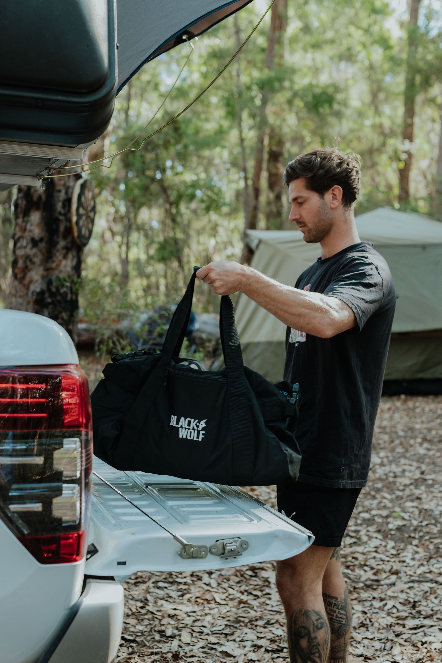 Man loading a Black Wolf duffel bag into a 4WD vehicle at a forest campsite.