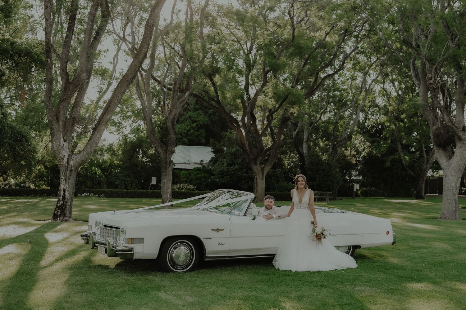 Bride in flowing white gown poses beside a vintage white convertible on a green lawn.