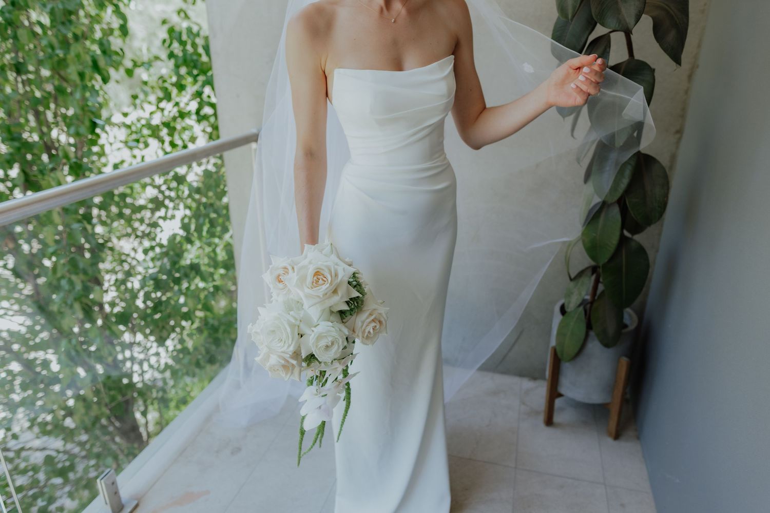 Elegant bride in strapless fitted white gown holding white floral bouquet on balcony with green trees.