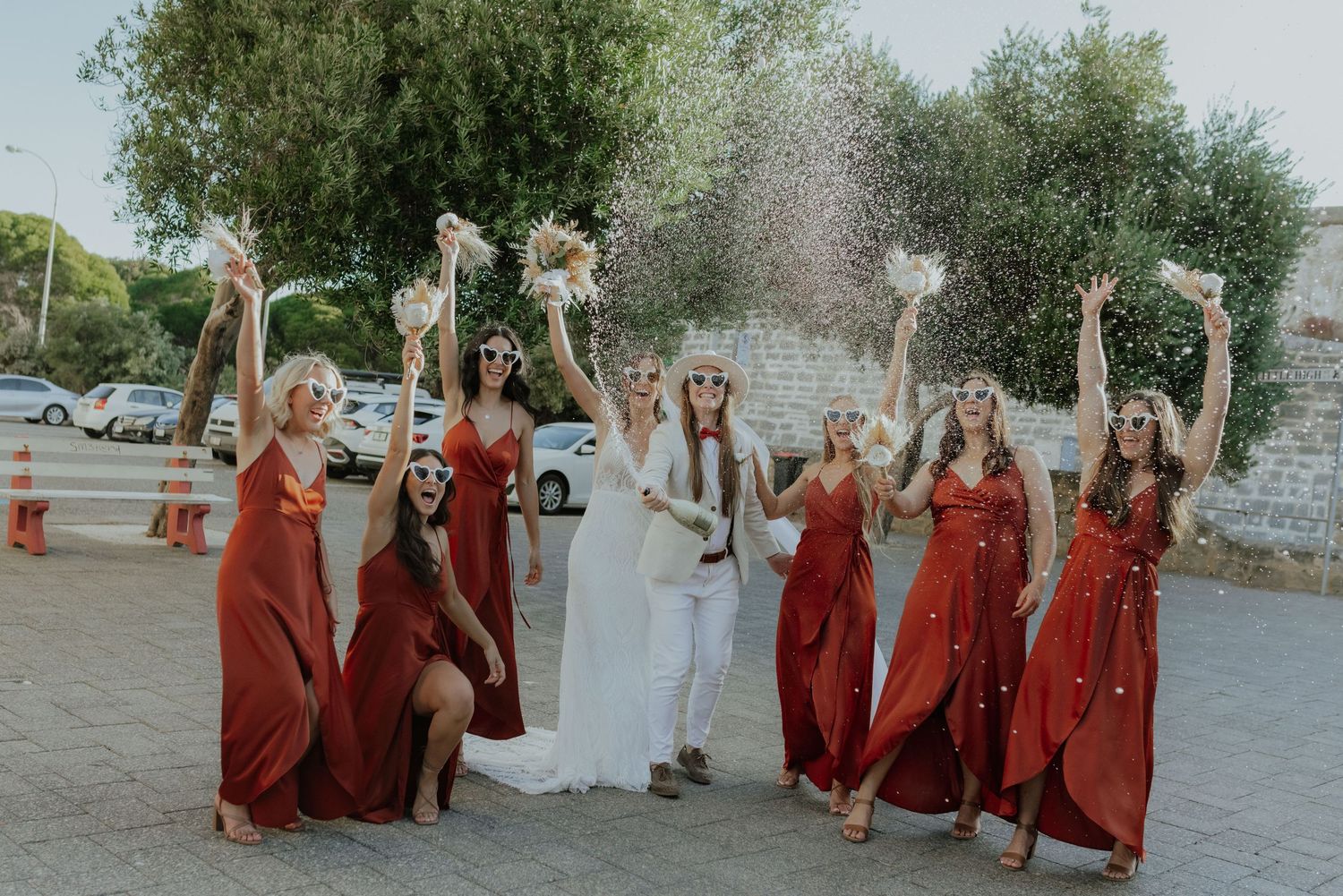 Bride with bridesmaids in red dresses toss bouquets joyfully outdoors on sunny wedding day.