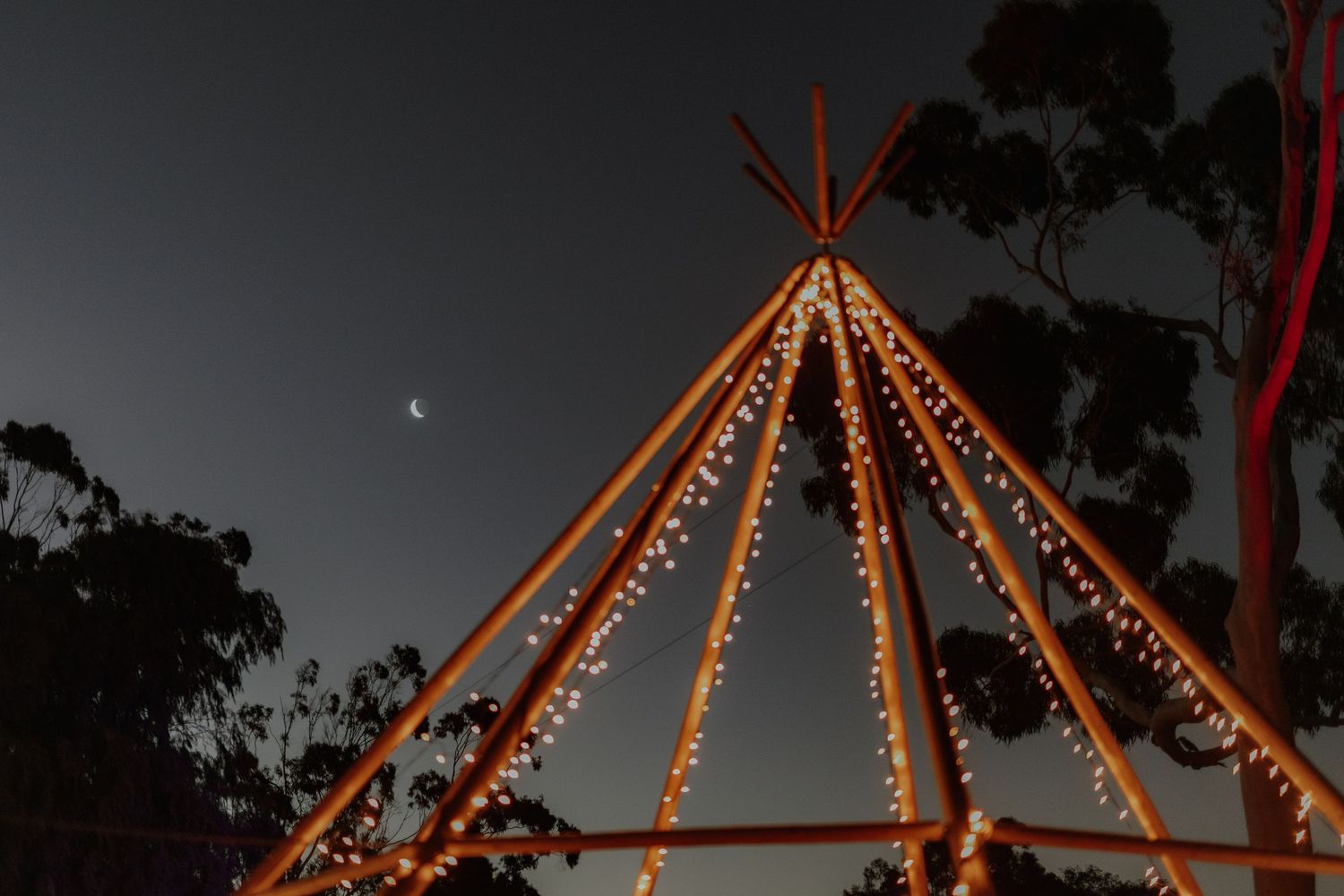 A teepee structure decorated with fairy lights glowing under a crescent moon at night.