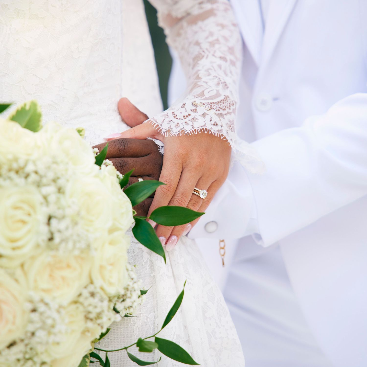 Interracial couple holding hands with wedding rings and a white rose bouquet.