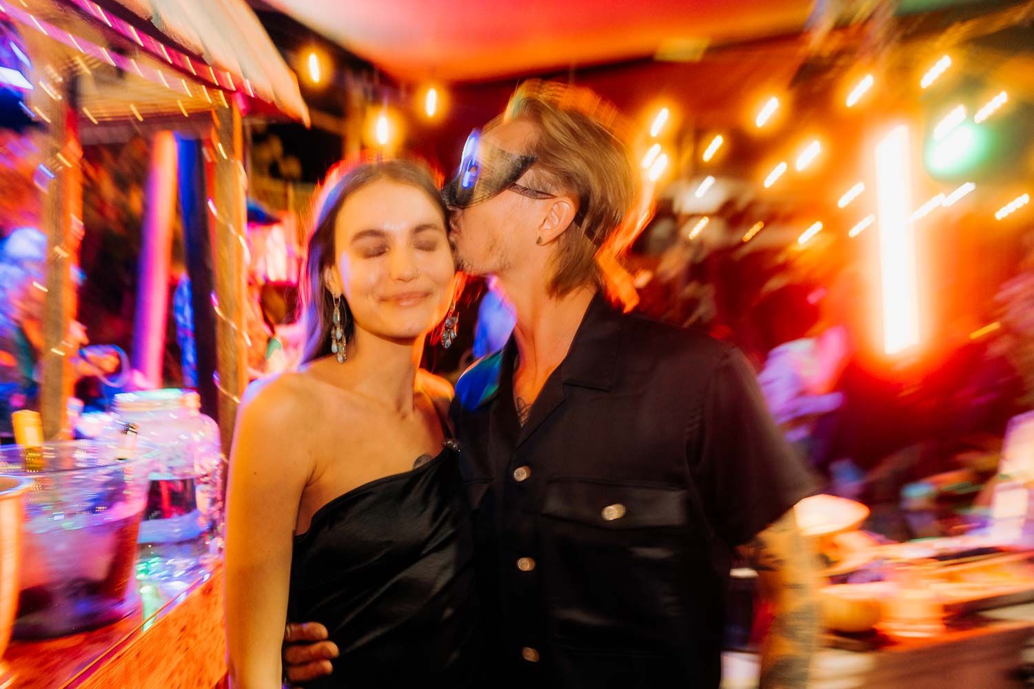 Couple at colorful nightclub event with bokeh lights, woman in black dress being kissed.