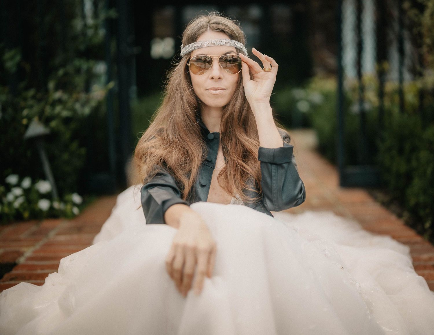 Woman in tulle skirt adjusting tinted sunglasses while sitting on brick pathway in garden.