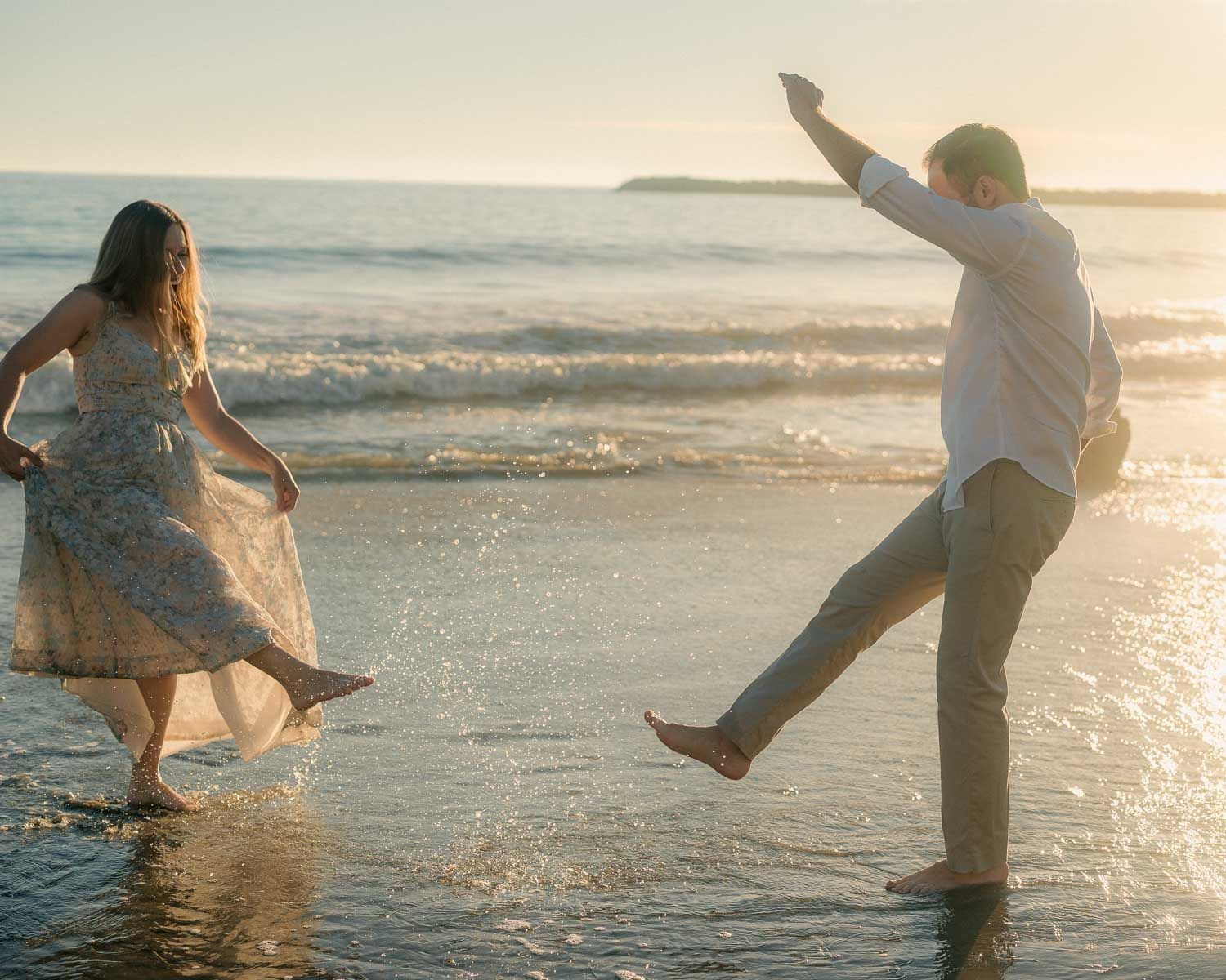 Playful couple splashing in ocean waves at sunset beach, laughing and kicking water.