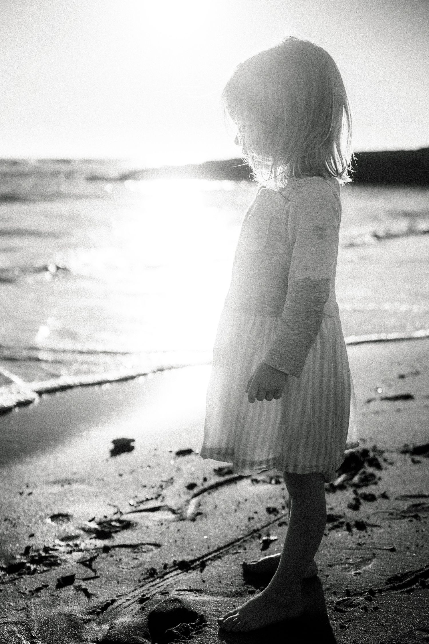 Young girl in white dress standing barefoot on a sunlit beach shore.