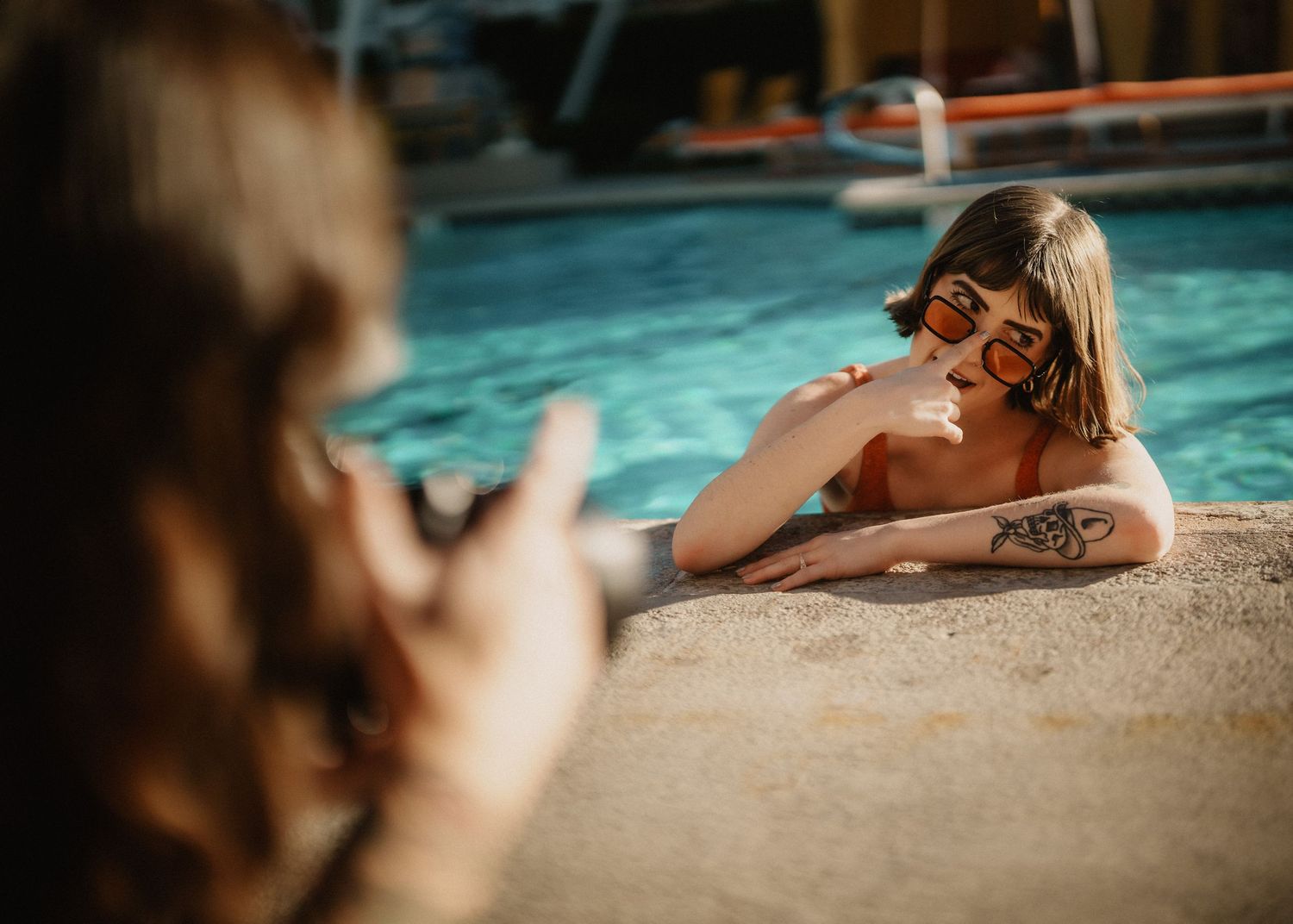 Tattooed woman in orange swimsuit smiling poolside while being photographed by another person.