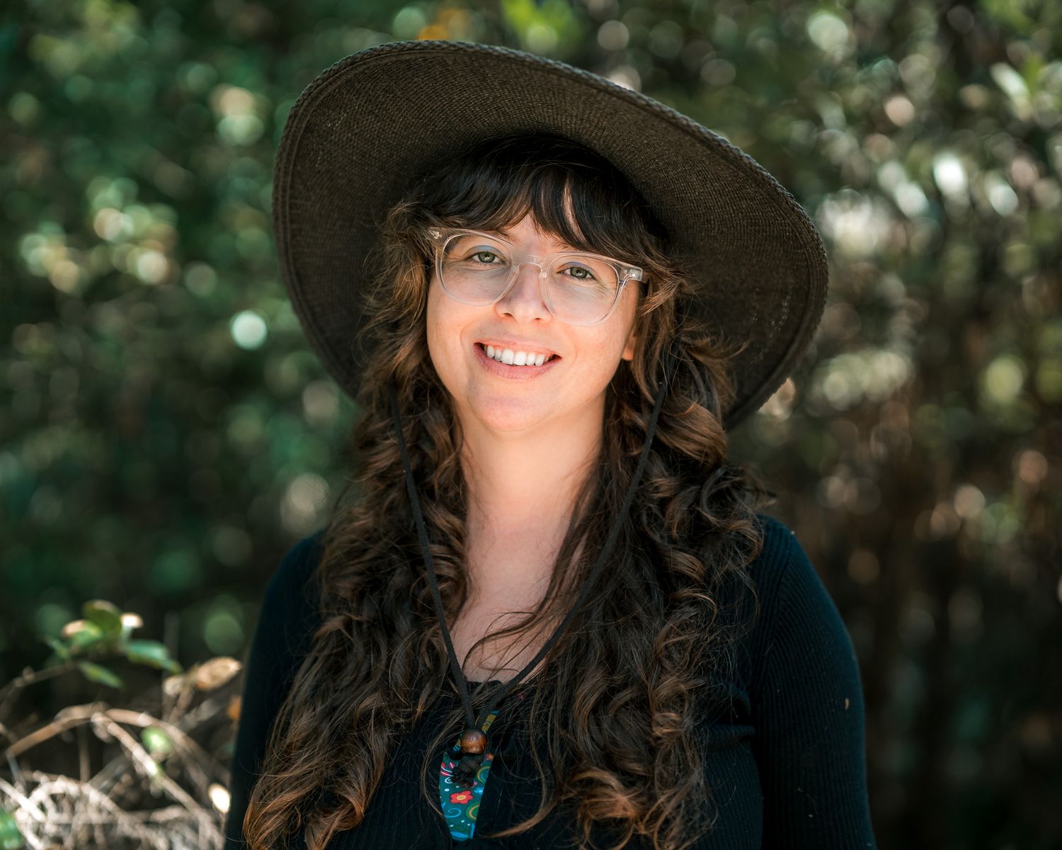 Smiling woman with curly hair wearing a wide-brim hat and turquoise necklace outdoors.