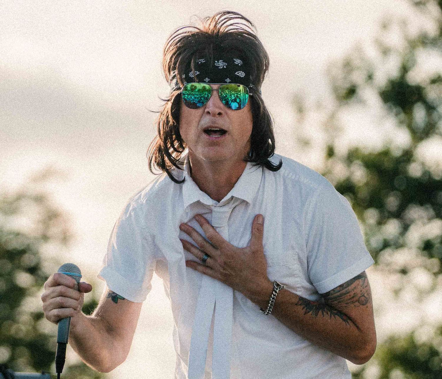 Male singer with bandana and mirrored sunglasses performs outdoors holding a microphone to his chest.