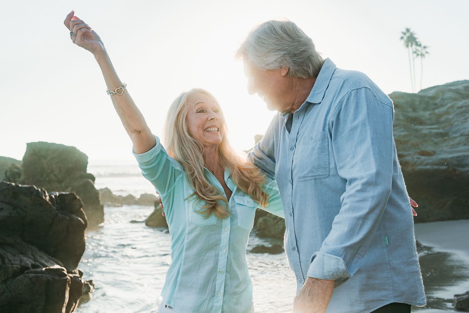 Joyful senior couple dancing on a rocky beach at golden hour, woman with arm raised celebrating by the ocean.