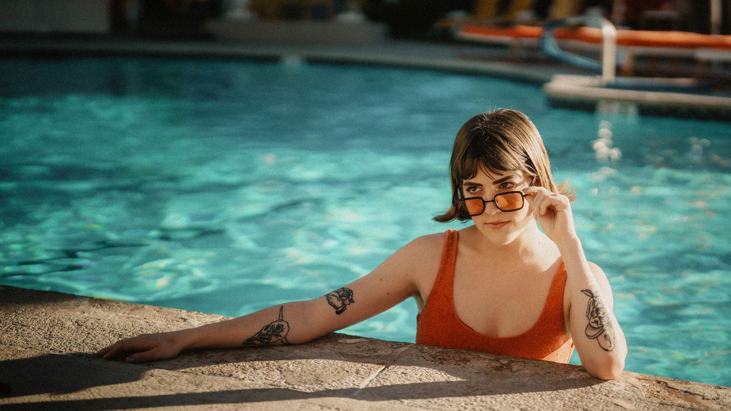 Smiling tattooed woman in orange swimsuit and sunglasses relaxing at the edge of a pool.