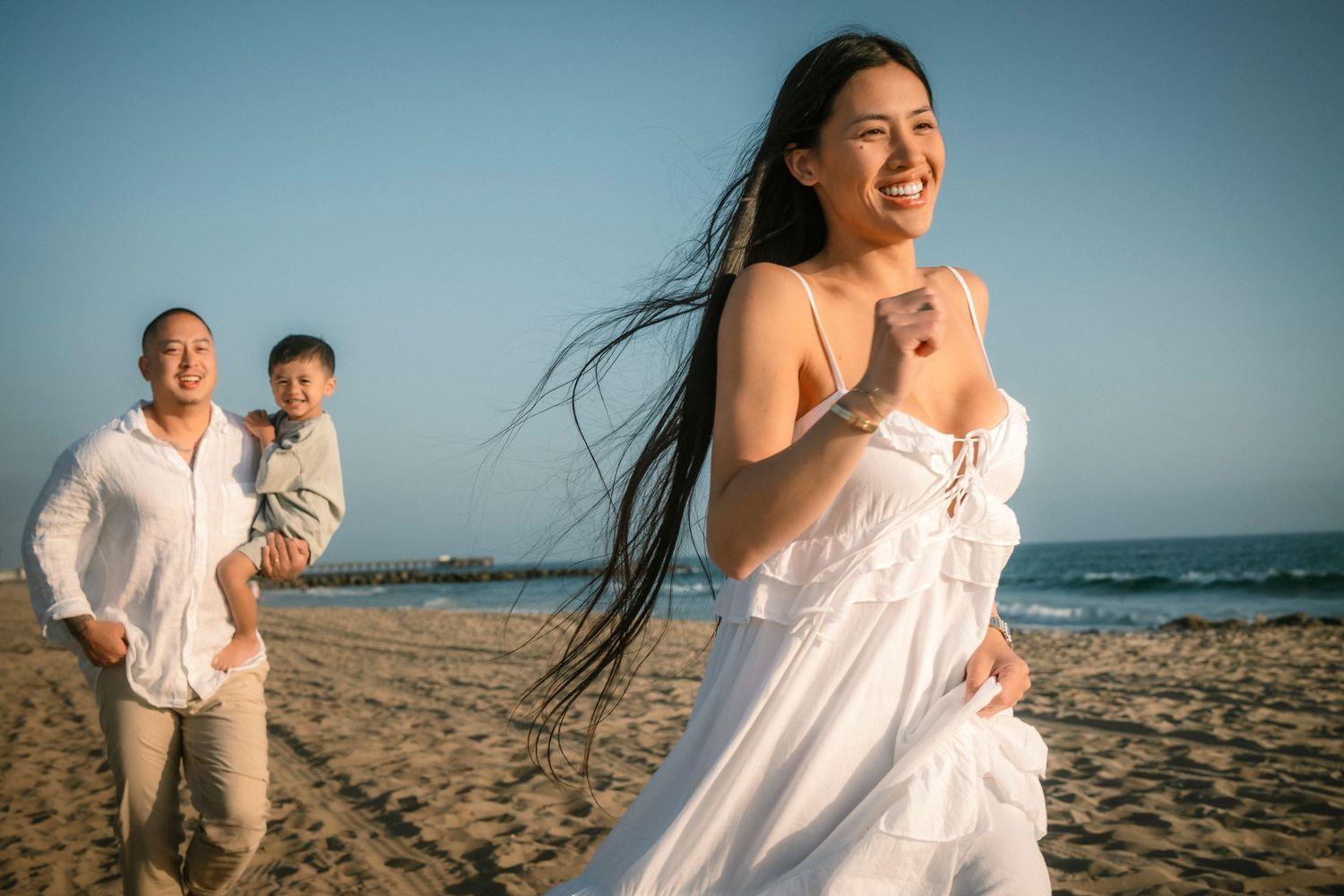 A happy Asian woman in a white dress laughs on the beach as her family follows behind her.