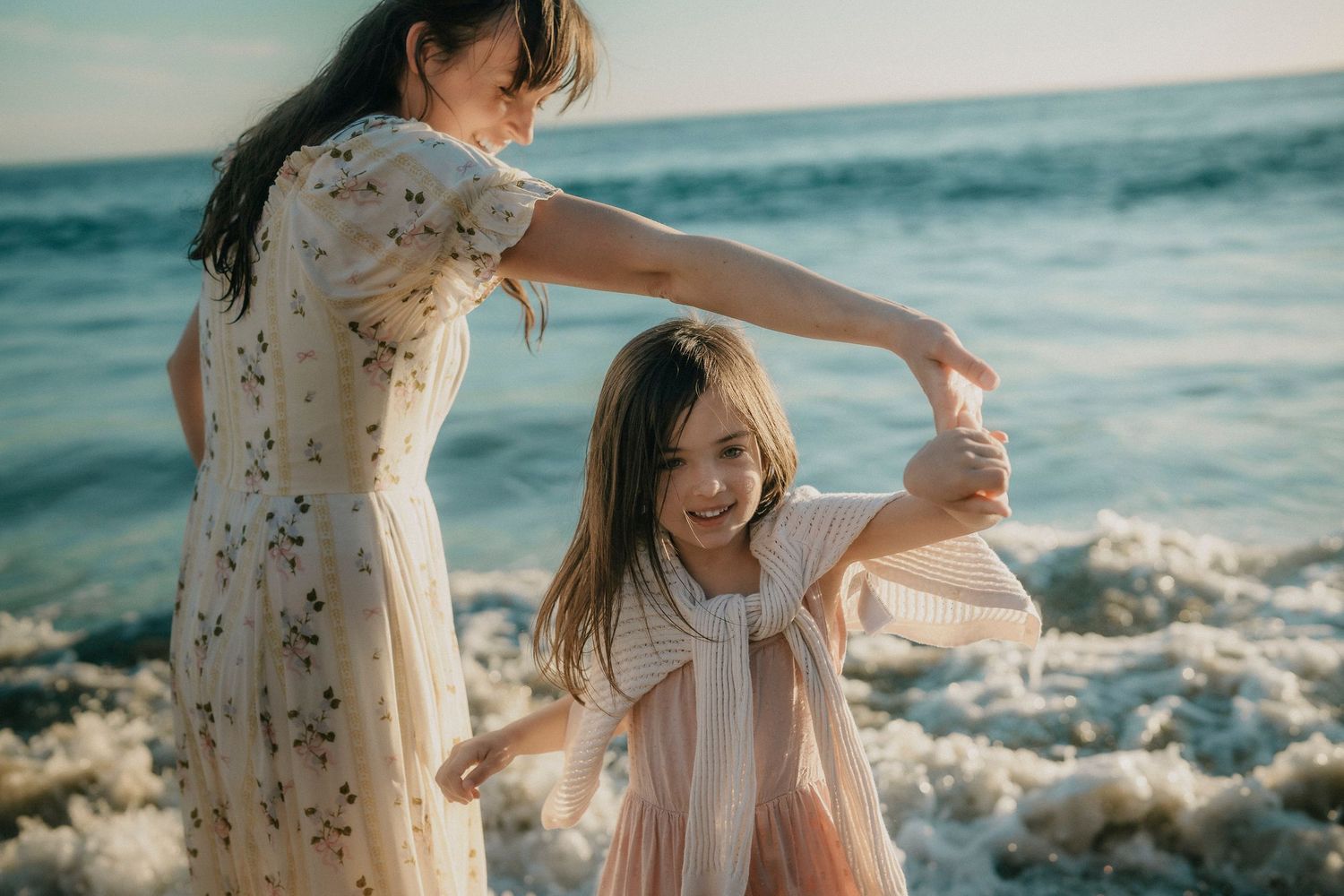 Mother spinning her laughing daughter on a beach with ocean waves splashing nearby.