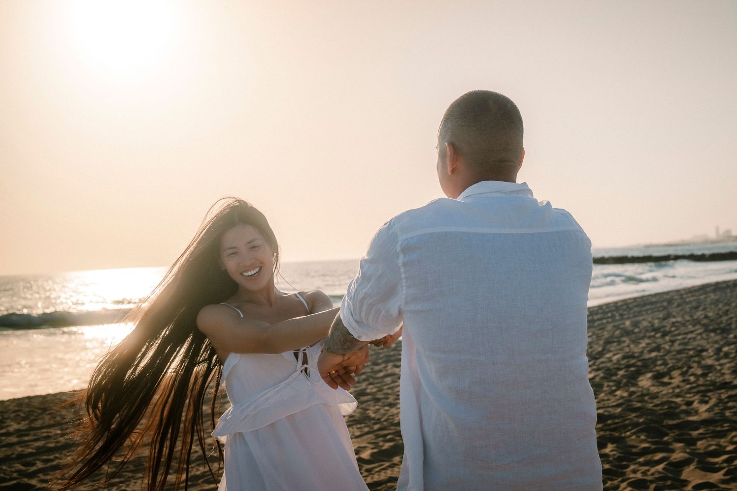 A laughing woman in a white dress spins while holding hands with a man on a sunny beach.