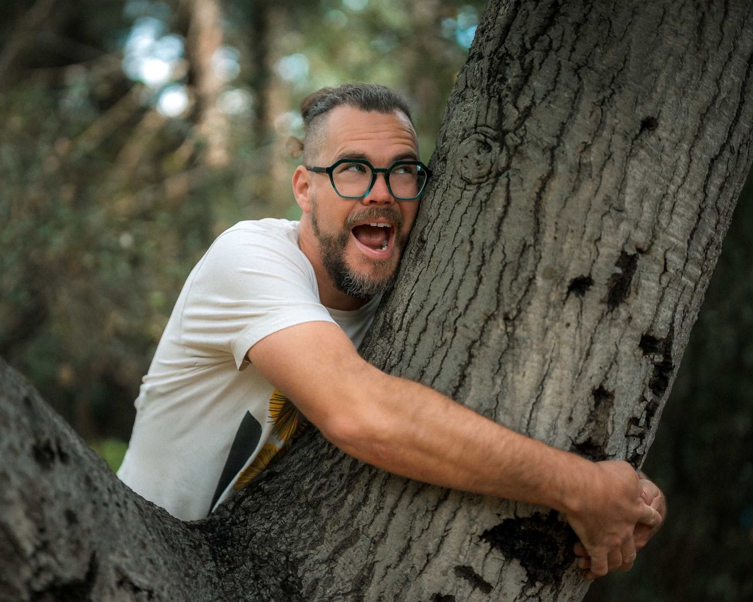 Bearded man with glasses hugs a large tree trunk with an expressive open-mouthed expression outdoors.