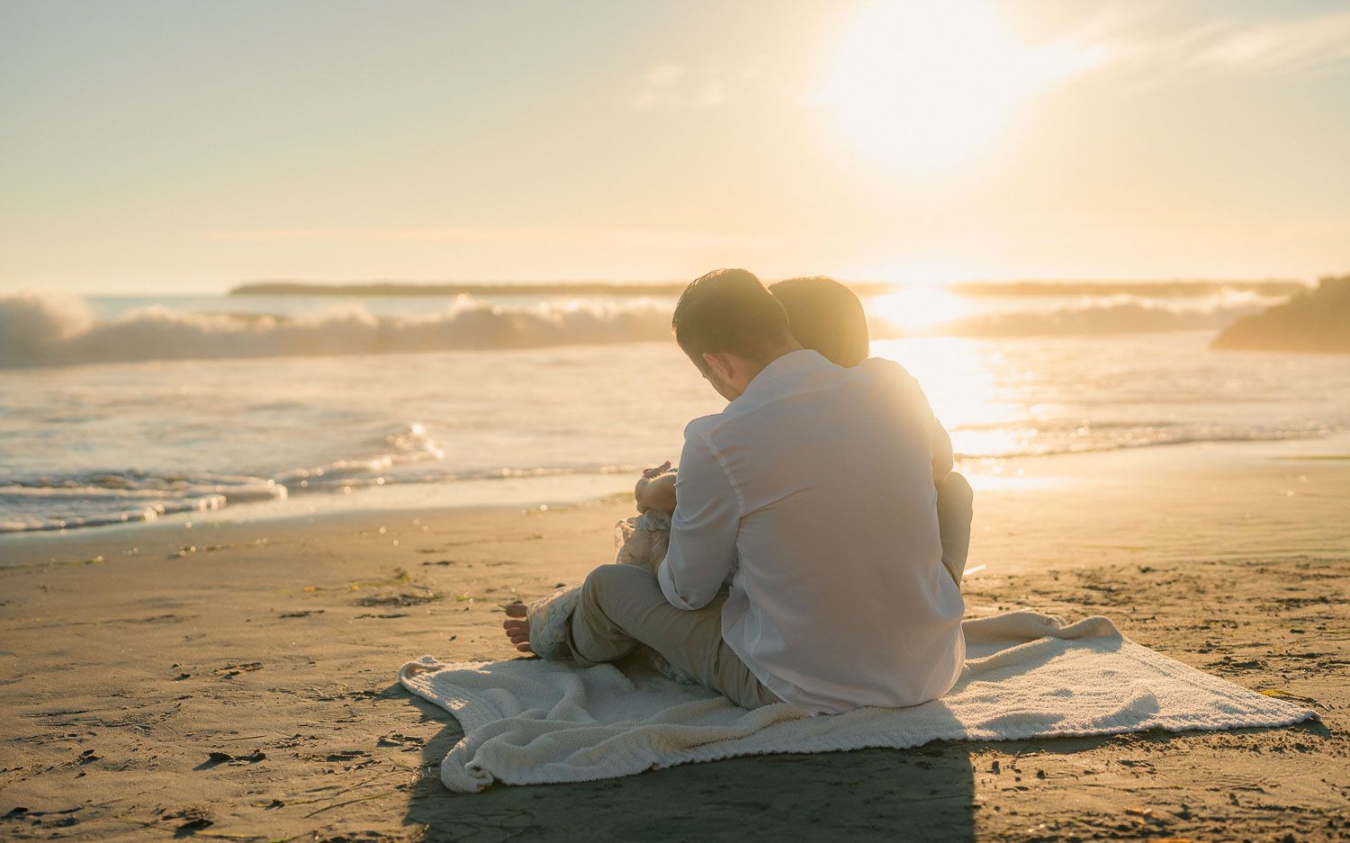Couple sitting together on beach blanket watching stunning golden sunset over ocean waves from behind.