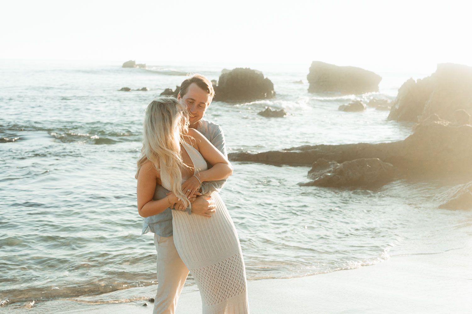 Young couple embracing romantically on a sunlit beach, woman in white lace dress with ocean rocks behind them.