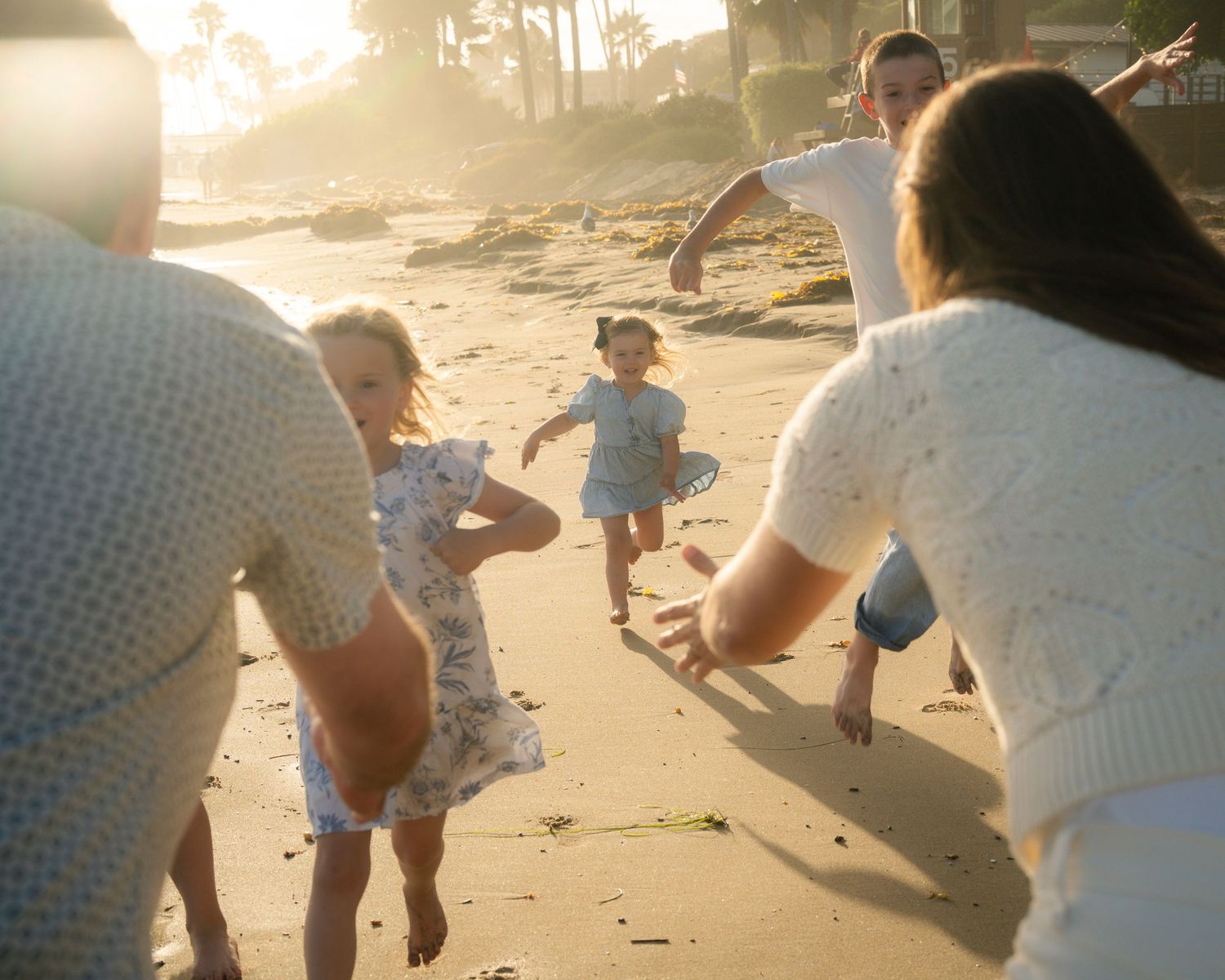 Two adults reach out to young children running toward them on a golden sunlit beach.