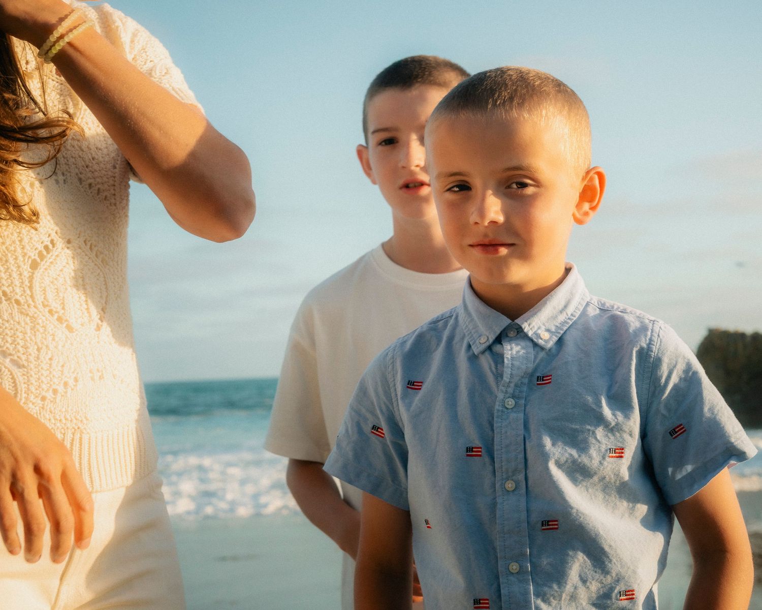 Two boys standing on a beach at golden hour, younger boy in blue shirt looking at camera.