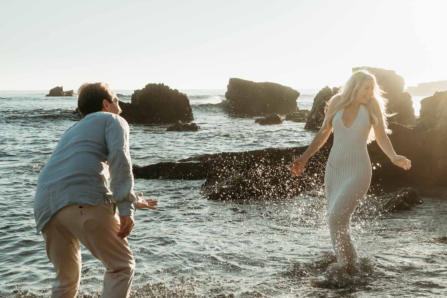 Playful couple splashing in ocean waves at sunset, woman in white dress laughing as water splashes around them.