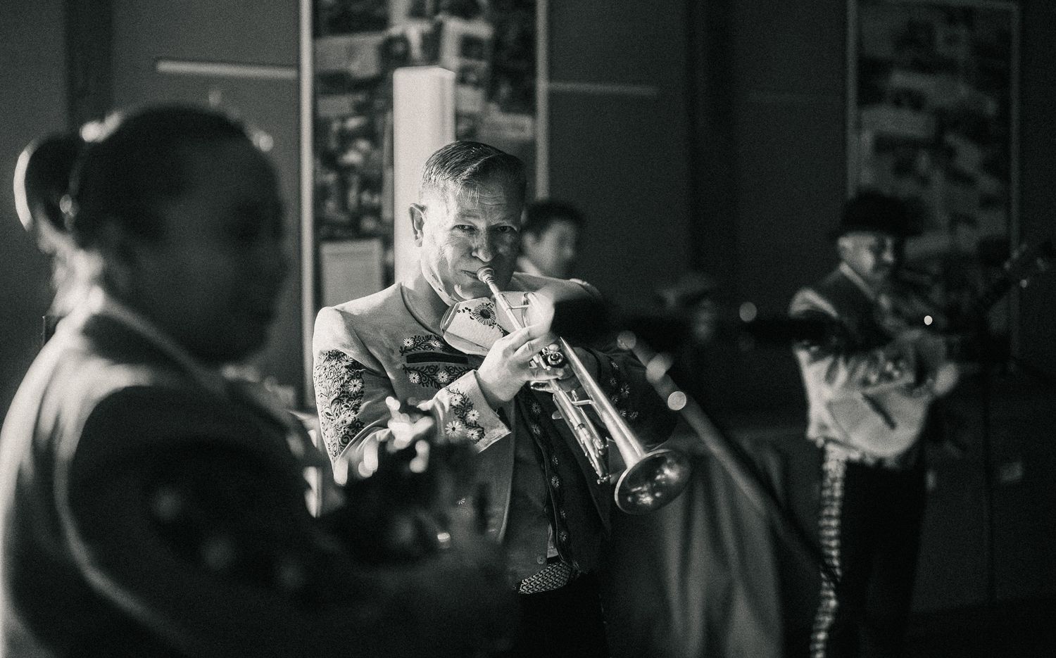 Black and white photo of mariachi musicians playing trumpets in a dimly lit backstage setting.
