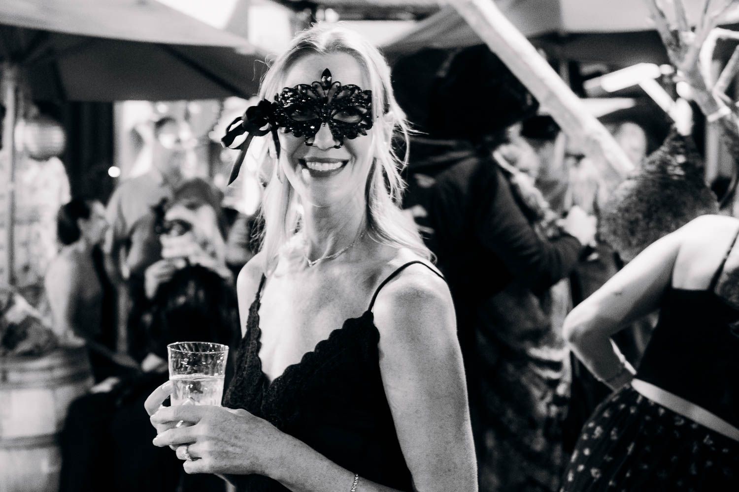 Black and white photo of a smiling woman in a masquerade mask holding a drink.