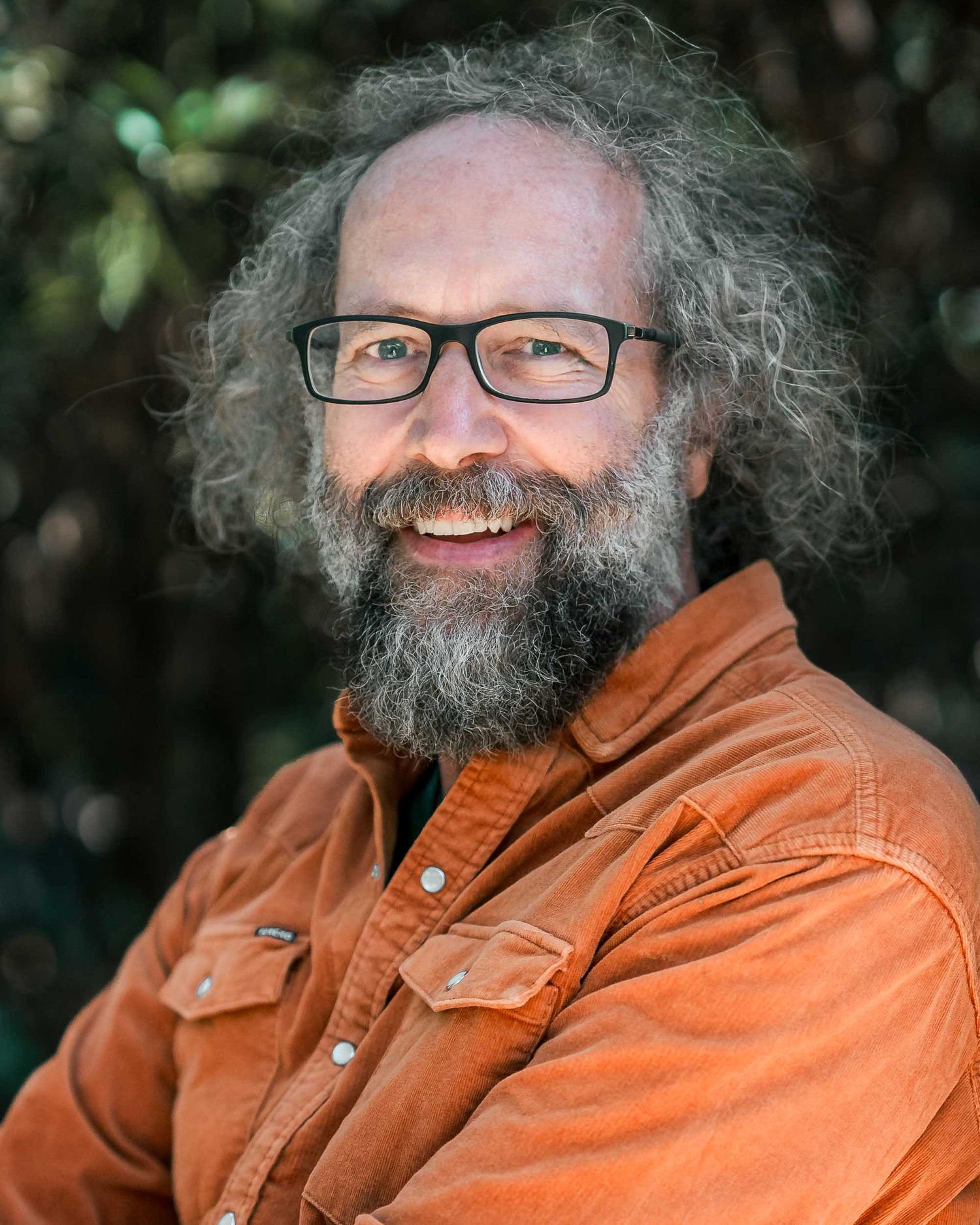 Smiling bearded man with curly hair and glasses wearing an orange shirt poses outdoors in nature.