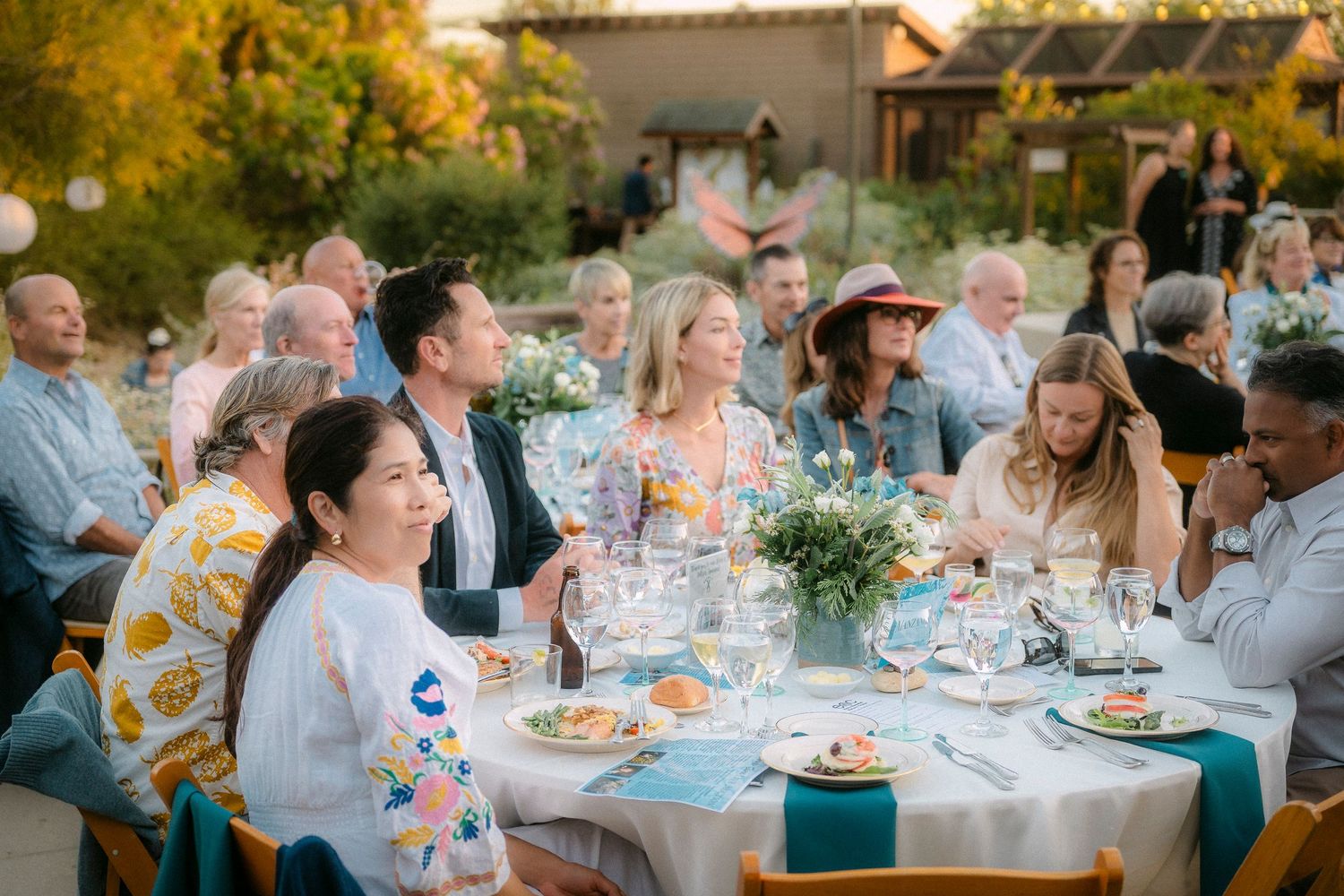 Guests enjoying an elegant outdoor dinner party with floral centerpieces and wine glasses at sunset.
