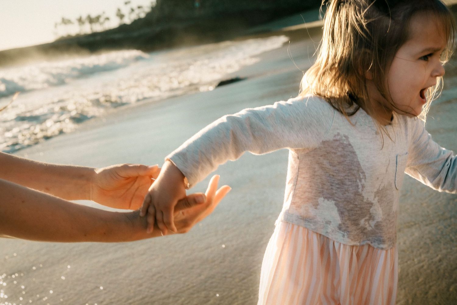 A young girl in a pink dress laughs while holding an adult's hand on a golden beach.
