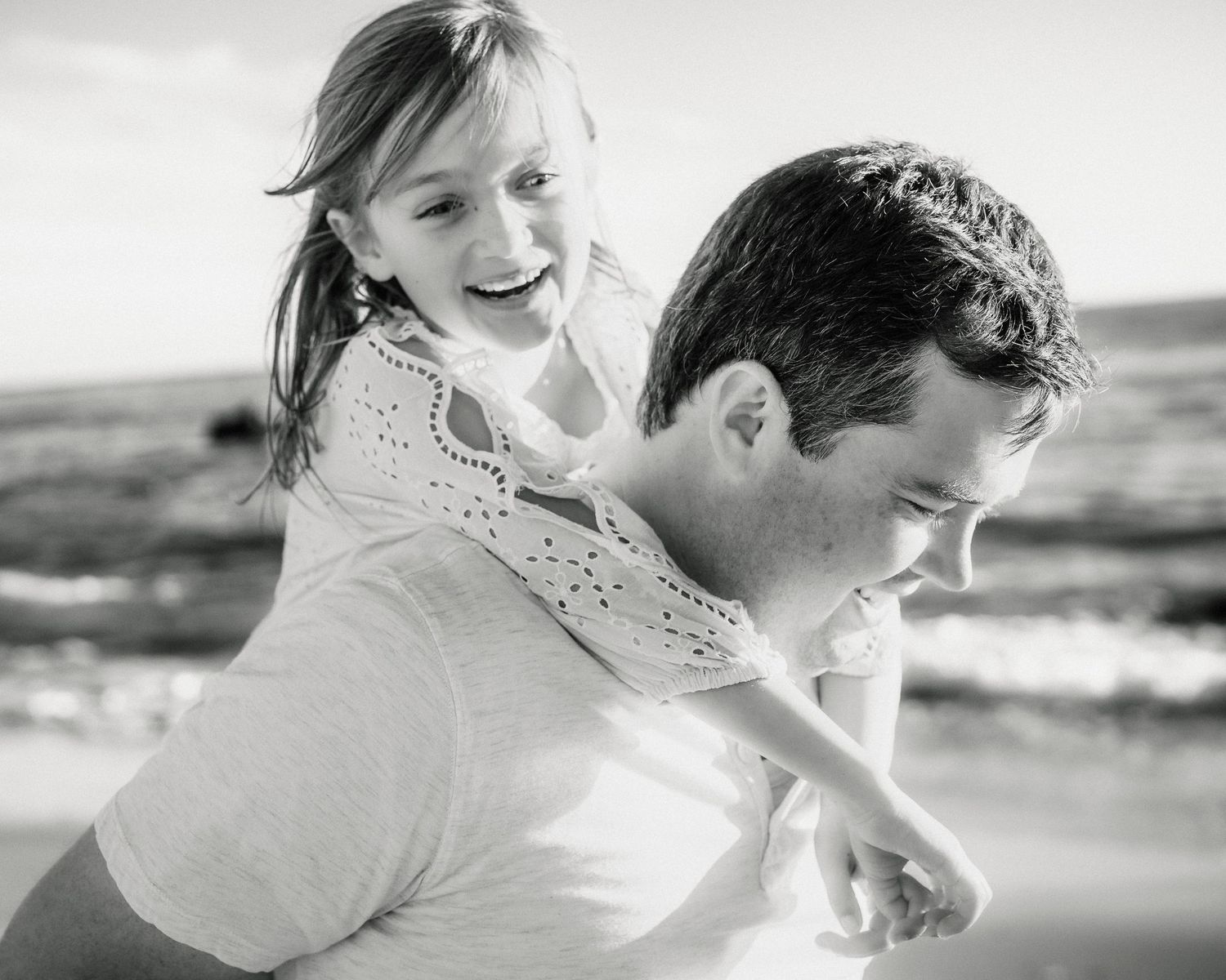 A black and white photo of a father giving his laughing daughter a piggyback ride on the beach.