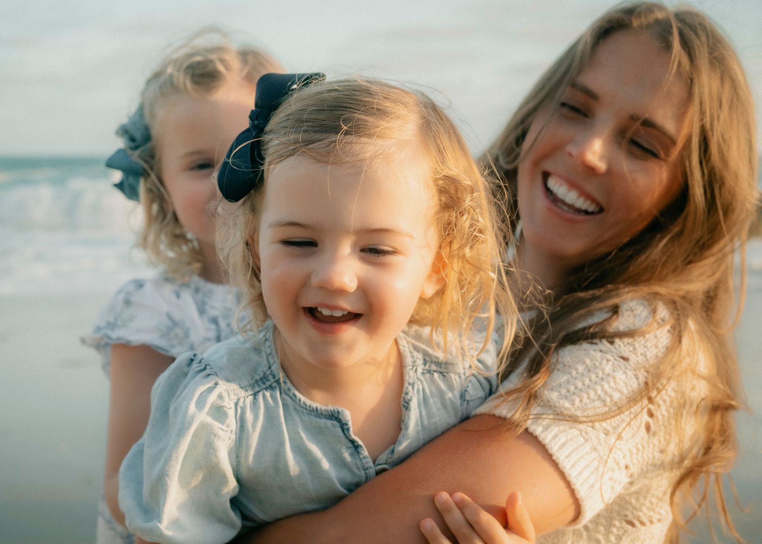Smiling mother hugging two young blonde daughters with hair bows at the beach.