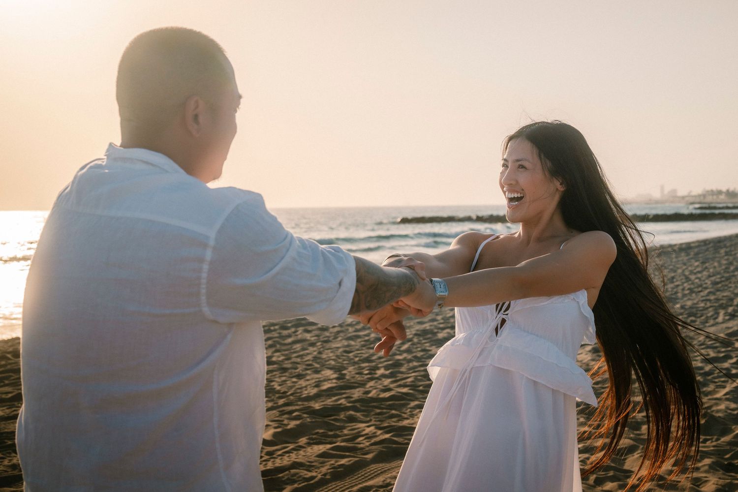 A couple holds hands and dances joyfully on a beach during a warm golden sunset.