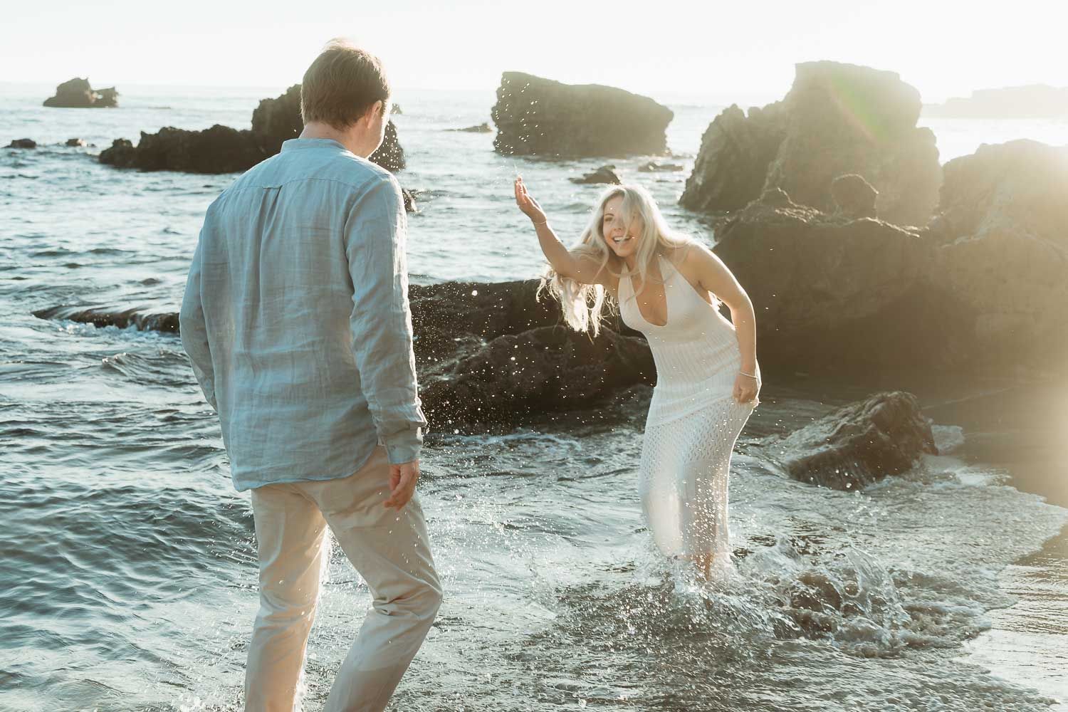 Couple laughing and splashing in ocean waves at sunset near rocky shore.