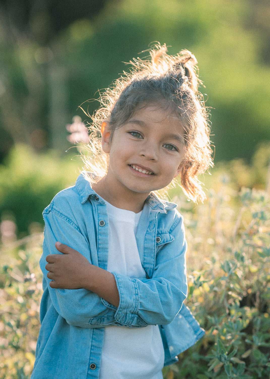 Smiling little girl in blue denim shirt with arms crossed standing in a sunlit field.