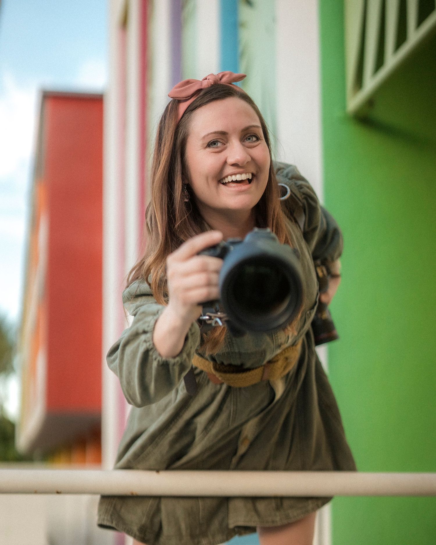Smiling woman holding camera toward viewer standing on colorful building balcony outdoors.
