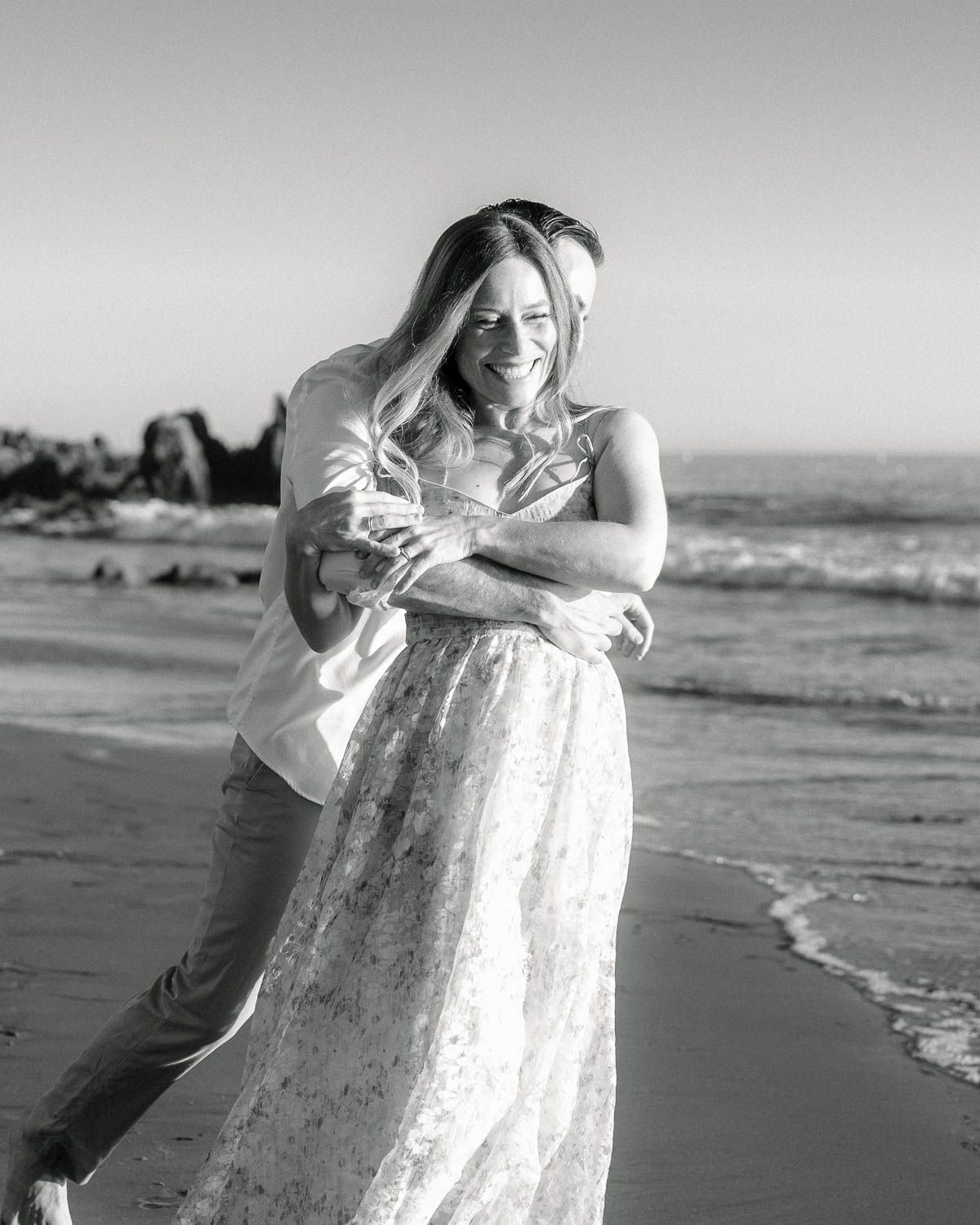 Black and white photo of couple embracing and laughing on beach with ocean waves.