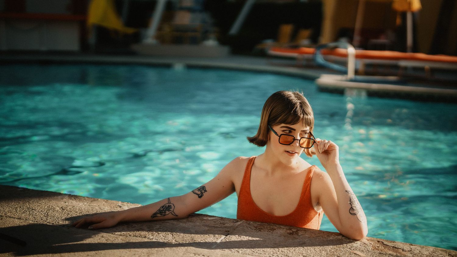 Woman in orange bikini with tattoos resting at pool edge wearing sunglasses in sunlight.