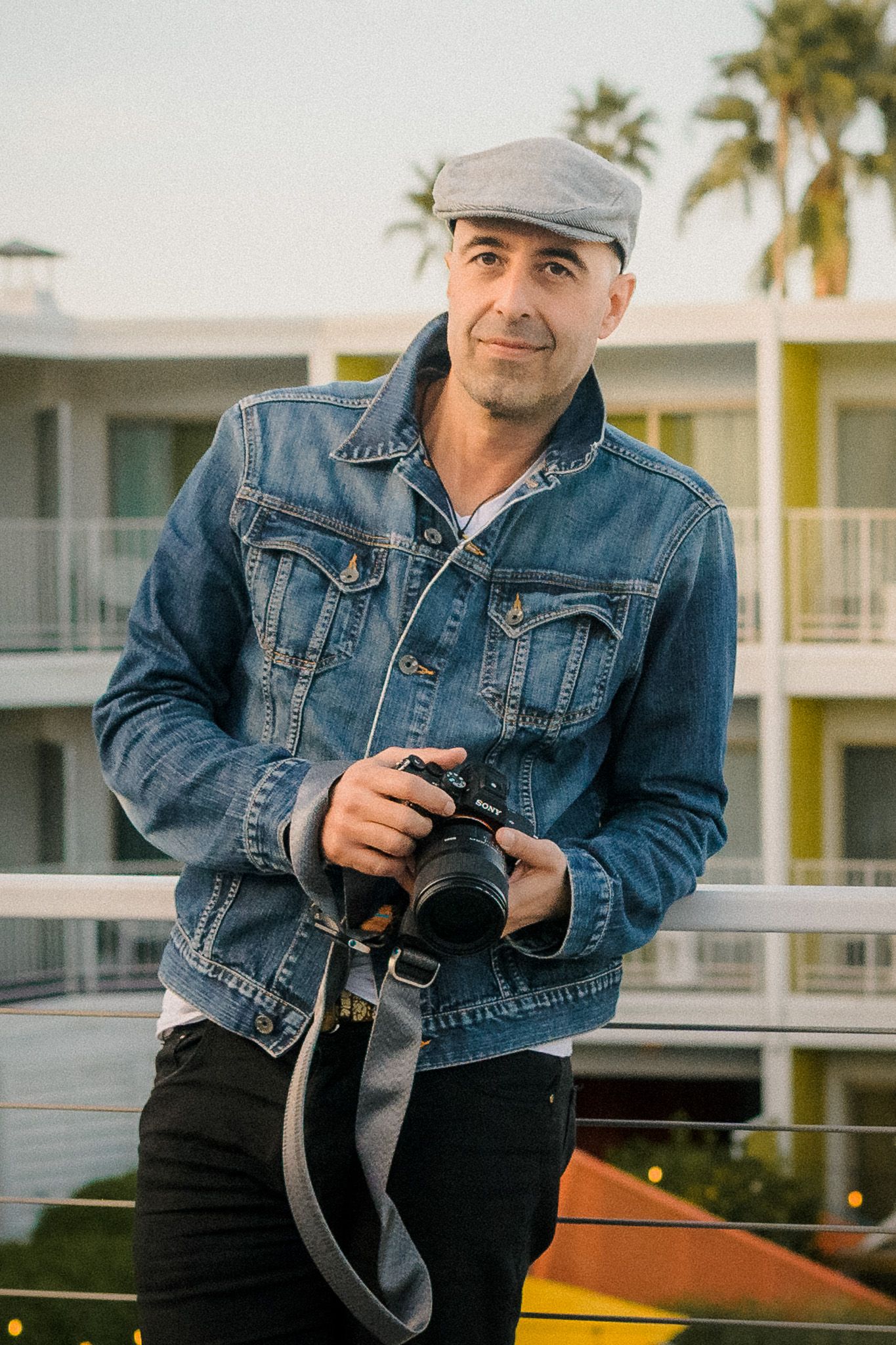 Male photographer in denim jacket and flat cap holding Sony camera at outdoor resort.