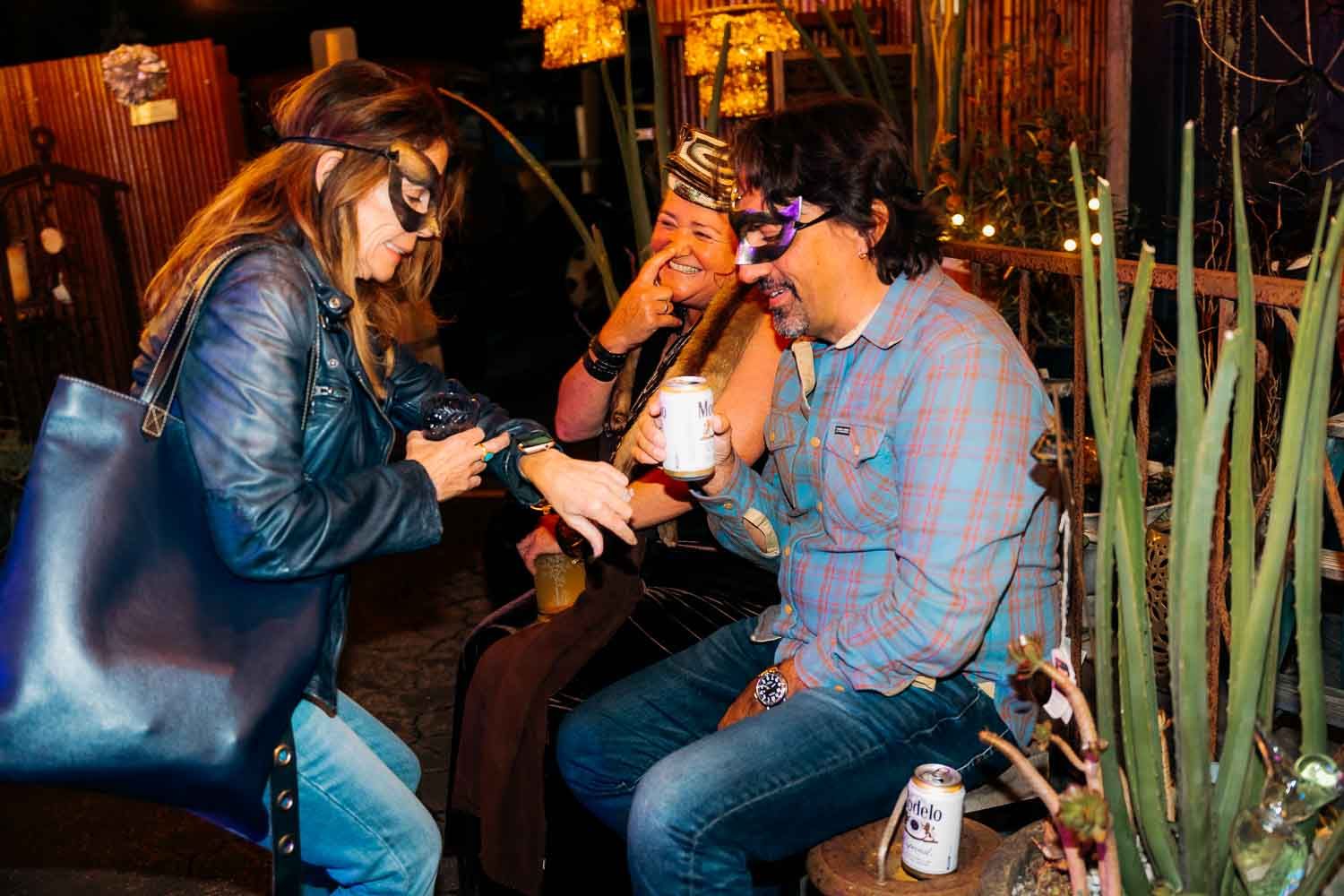 Three friends laugh and socialize outdoors at a casual evening gathering with drinks and string lights.