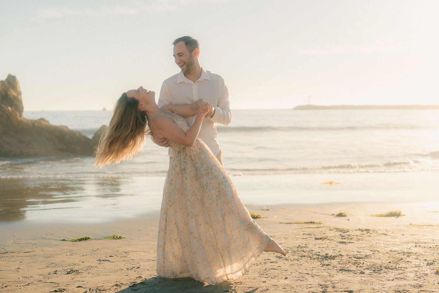 Man dipping woman in elegant lace dress on sunlit beach with rocky coastline backdrop.